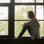 Thoughtful girl sitting on sill embracing knees looking at window