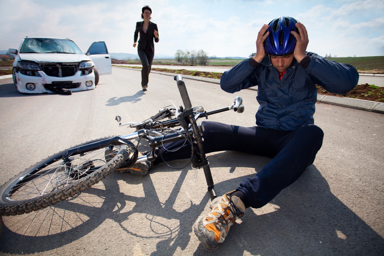 Bicyclists injured in a car accident lying down on the road and holding his head with helmet. A person who is driving car running to helps to injured man.