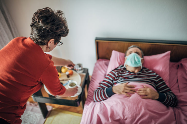 Carer giving tea to patient in bed with mask on