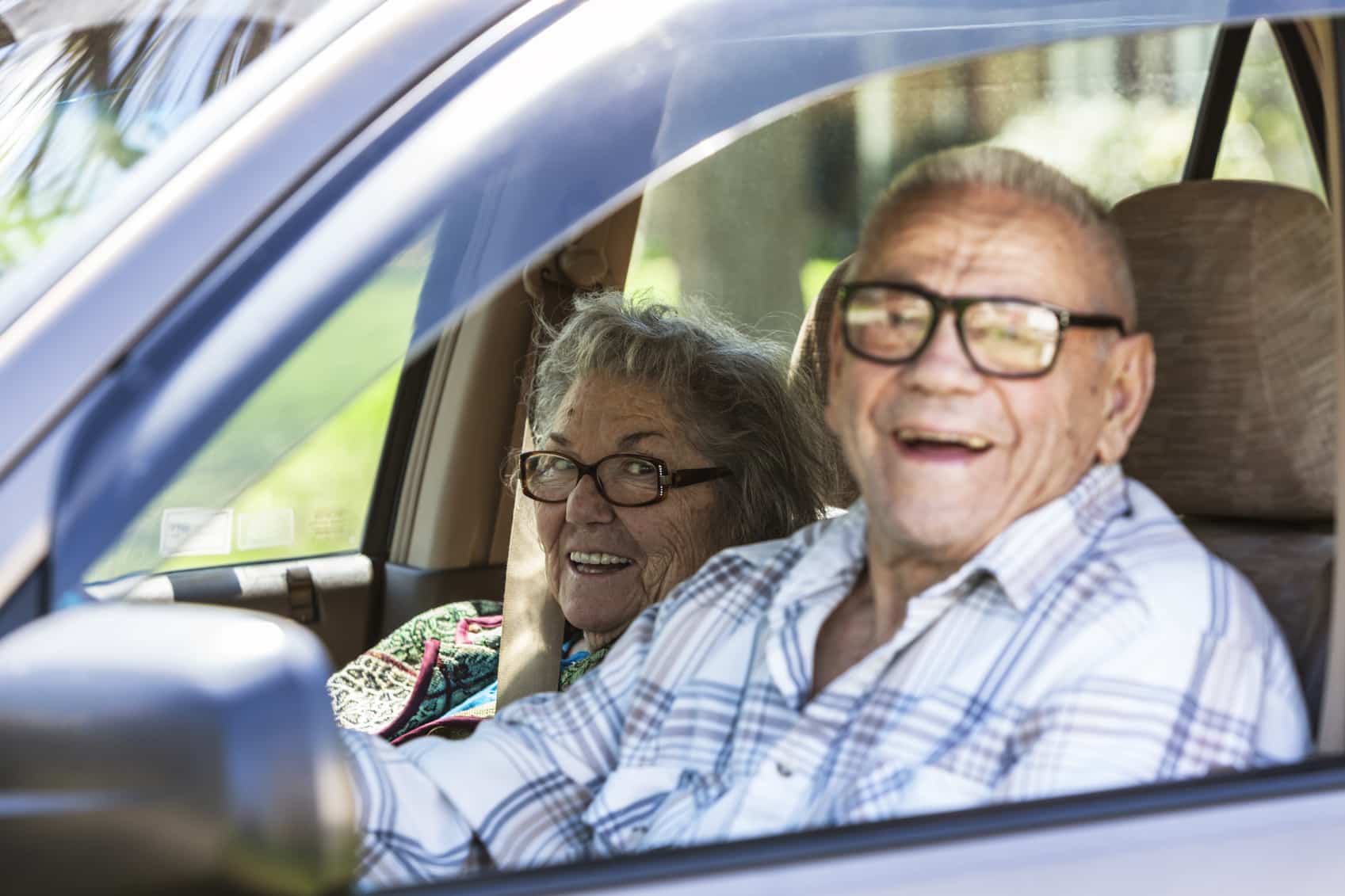 Elderly senior adult married heterosexual couple - 89 and 87 years old - laughing at the camera as they drive past in their trusty, well-worn car. Focus on the woman.